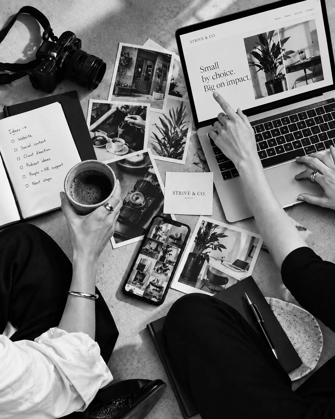 Hands working over a Strive & Co. desk — camera, coffee, printed photographs, and a laptop reading Small by choice. Big on impact.